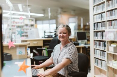 A librarian with a welcoming smile and hair pulled back, sits behind the circulation desk. The librarians arms reach forward with one hand resting on a computer keyboard and the other hand resting on a computer mouse.